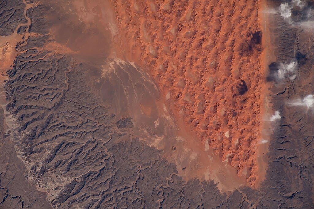 International Space Station photo of Algeria’s Sahara showing red sand dunes meeting dark rocky desert terrain.