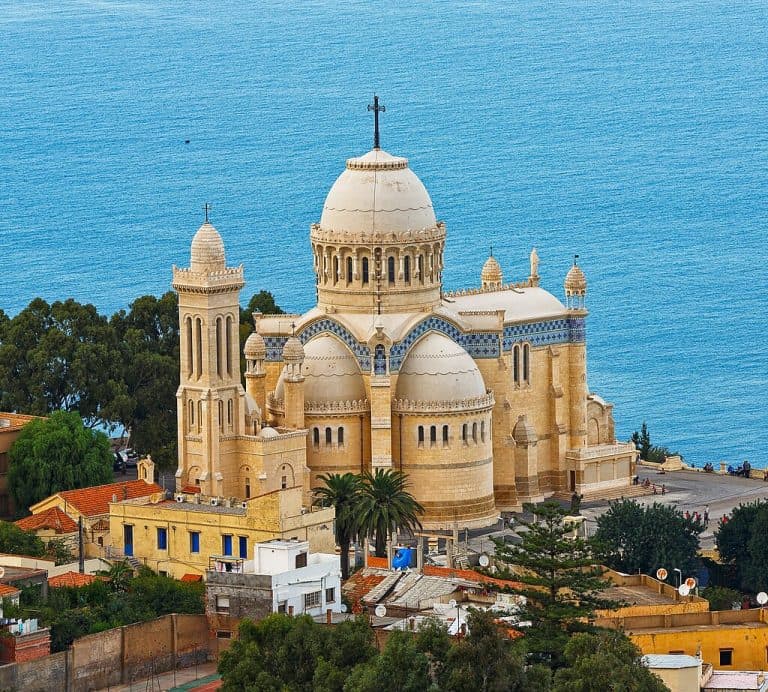 Elevated view of Notre-Dame d’Afrique Basilica in Algiers, with domes and tower in the foreground and the Mediterranean Sea behind.