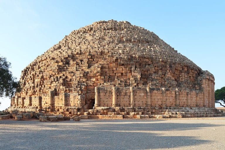 Circular stone mausoleum on a hilltop in Algeria, with a ring of columns and a stepped conical roof rising above the surrounding landscape.