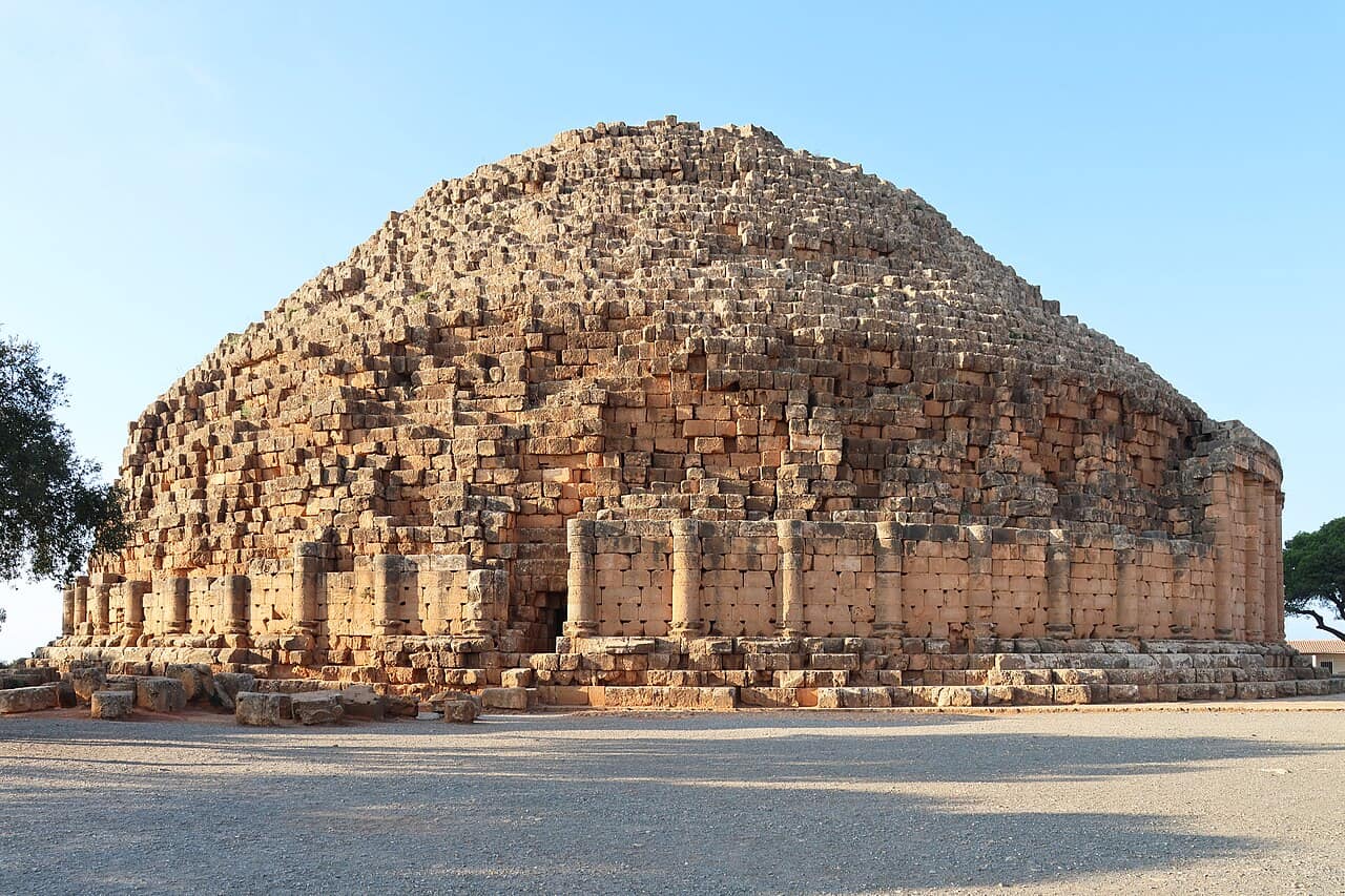 Circular stone mausoleum on a hilltop in Algeria, with a ring of columns and a stepped conical roof rising above the surrounding landscape.