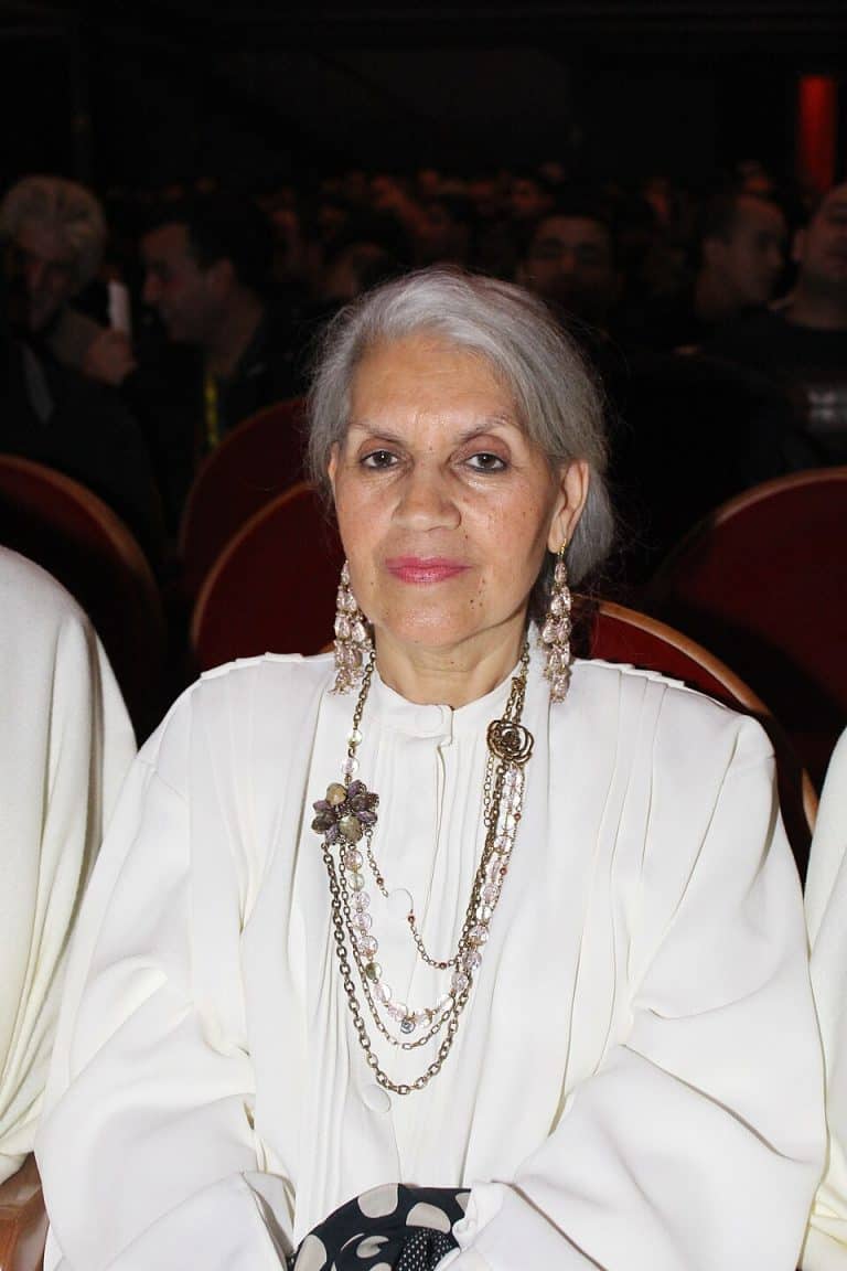 Houria Aïchi seated in an auditorium, wearing a white blouse and layered necklaces.