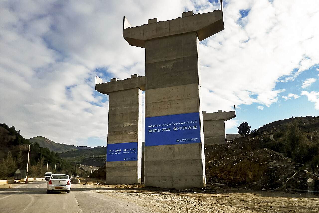 Unfinished concrete bridge supports beside a road near El Hamdania, Algeria, with signs in Arabic, Chinese and French.
